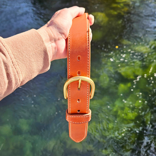 Sand Creek chestnut leather belt with brass D buckle held above a creek